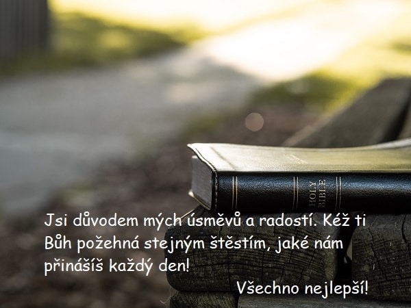 Náboženská gratulace k narozeninám na pozadí bible položené na dřevěné lavičce v přírodě u cesty.