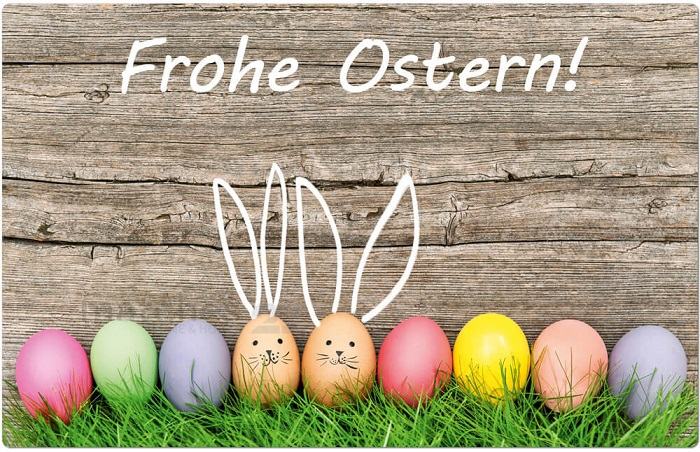 Řada barevných vajíček s obličeji zajíčků s ušima na dřevěném pozadí s nápisem Frohe Ostern!