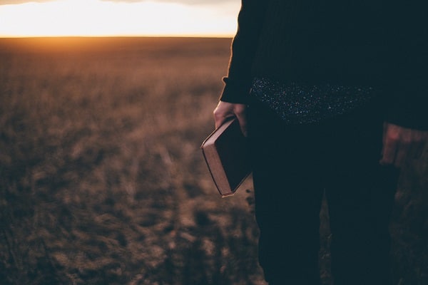 Osoba stojící na louce s biblí v ruce při západu slunce.